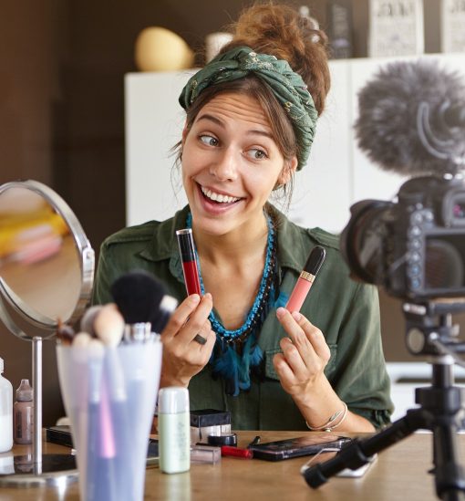 Portrait of excited cheerful young woman vlogger holding two lipsticks of different brands and compa