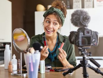 Portrait of excited cheerful young woman vlogger holding two lipsticks of different brands and compa